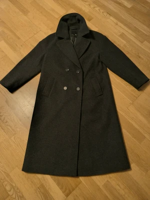 Mörkgrå lång kappa från 157 - Stilren mörkgrå kappa från 157 med dubbelknäppning och klassisk krage. Lång modell med två fickor framtill och helfodrad insida. Perfekt för dig som vill ha en tidlös och snygg look under kyliga dagar.