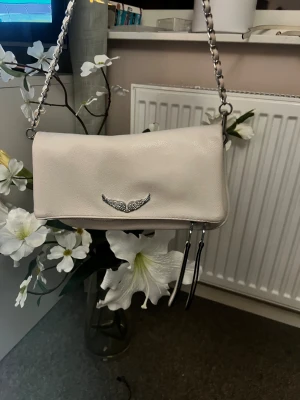 Vit axelväska med kedjerem - Snygg vit axelväska i skinnimitation med silverfärgad kedjerem och dekorativt emblem framtill. Väskan har dragkedja baktill och två långa tofsar som detalj. Perfekt storlek för det viktigaste och en stilren look. Jag vet inte om väskan är äkta det är därför det är så lågt pris. Jag fick den av en vän som hade klippt bort etiketten. 