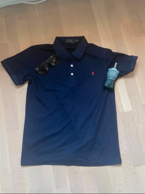 Mörkblå piké från Polo Ralph Lauren - Snygg mörkblå pikétröja från Polo Ralph Lauren i slim fit-modell. Klassisk krage, tre vita knappar och röd broderad logga på bröstet. Materialet är mjuk bomull som känns skönt mot huden. Perfekt för dig som gillar stilrena och tidlösa plagg.