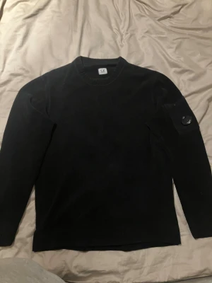 Svart stickad tröja från C.P. Company - Svart stickad tröja från C.P. Company med rund hals och lång ärm. På vänster ärm finns en ficka med knapp och den ikoniska C.P. Company-linsen. Tröjan har en clean och stilren look, perfekt för dig som gillar kvalitativt material och snygga detaljer. Passar storlek S, äkta och aldrig använd. Rätt så limiterad. Kan diskutera priset något.