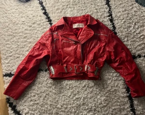 Röd skinnjacka från Epiderme - Säljer en röd skinnjacka från Epiderme med coola silvriga detaljer och bälte i midjan. Jackan är croppad, har dragkedjor och flera fickor framtill. Perfekt för dig som vill sticka ut med en edgy stil