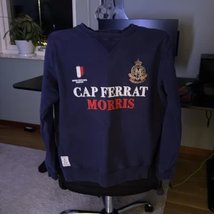 Mörkblå sweatshirt Morris XS - Mörkblå sweatshirt från Morris i storlek XS med broderade detaljer och tryck på bröstet samt en röd siffra 6 på ena ärmen. Tröjan har rund hals, ribbade muddar och mjukt bomullsmaterial. Snyggt broderad emblem och texten CAP FERRAT MORRIS framtill.