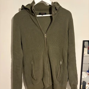 Olivgrön stickad hoodie från Indicode - Säljer en olivgrön stickad hoodie från Indicode som är extremt snygg och eftertraktad!, med dragkedja framtill och två fickor med dragkedja. Tröjan har en quiltad huva i blankt material och långa ärmar. Perfekt för dig som gillar chill och stilrena plagg 