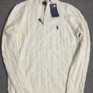  Polo Ralph Lauren - Kabelstickad vit tröja från Polo Ralph Lauren med half zip och broderad logga på bröstet. Tröjan har ribbad krage, muddar och nederkant samt långärmad passform. Perfekt för dig som gillar klassisk och clean stil. En helt ny, kan diskutera priset!