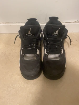 Svarta Nike Air Jordan 4 sneakers - Säljer ett par svarta Nike Air Jordan 4 sneakers med vita detaljer och meshpaneler. Skorna har snörning, hög siluett och den klassiska Jumpman-loggan på plösen och hälen. Materialet är en mix av skinn och syntet, med platt sula och rund tå. Perfekt för dig som gillar streetwear.