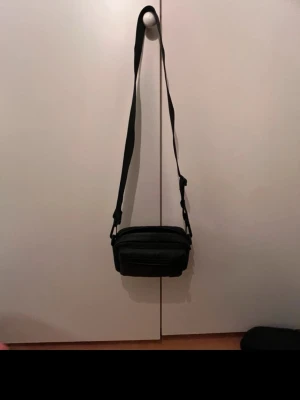 Svart axelväska med dragkedja - En stilren svart axelväska med justerbar rem och flera fack framtill. Väskan har en enkel, rektangulär form och stängs med dragkedja. Perfekt för att bära med sig det viktigaste och passar till många olika outfits.