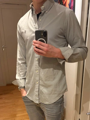  Skjorta från Polo Ralph Lauren - Snygg grå skjorta från Polo Ralph Lauren med klassisk button-down krage och broderad logga på bröstet. Custom fit-modell i mjuk bomull, perfekt för dig som gillar stilren och enkel design. Långärmad med vita knappar framtill.
