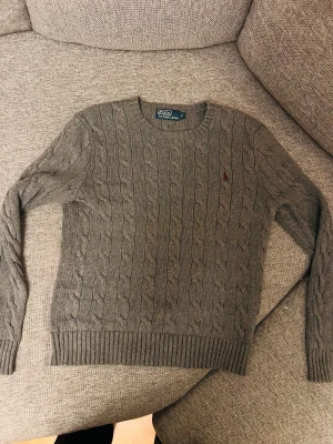 Polo Ralph Lauren - Säljer en grå kabelstickad tröja från Polo Ralph Lauren i storlek S. Tröjan har rund hals, långa ärmar och ett rött broderat logomärke på bröstet. Materialet är mjukt och stickat, perfekt för kyliga dagar. Ribbstickade muddar vid ärmslut och nederkant.