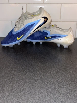 Nike Phantom 6 Elite - Nike Phantom fotbollsskor i blått och vitt med ljusblå detaljer och gula swoosh-loggor. Skorna har snörning, platt sula med dobbar och en rund tå. Ovandelen är tillverkad i syntetmaterial med stickad textil för extra komfort och passform. Perfekta för dig som vill sticka ut på planen. Använda 2-3 gånger och har inga skador alls! 