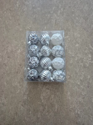 Julkulor - Säljer ett paket med 12 julgranskulor i silver och vitt från Rusta. Kulorna är ca 3,5 cm i diameter och har olika mönster – några är glittriga, andra har räfflad eller facettslipad yta. Perfekta för att ge granen en frostad och lyxig vibe.