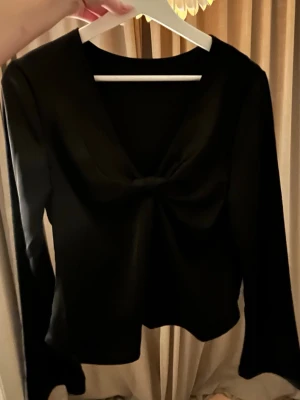 Svart blus med v-ringning - Säljer en svart blus med v-ringning och snygg detalj framtill. Blusen har långa ärmar med lätt utsvängda avslut och är gjord i ett glansigt, mjukt material som ger en lyxig känsla. Perfekt att styla med jeans eller kjol för en trendig look. Storleken är M men jag skulle säga att den sitter mer som en S.