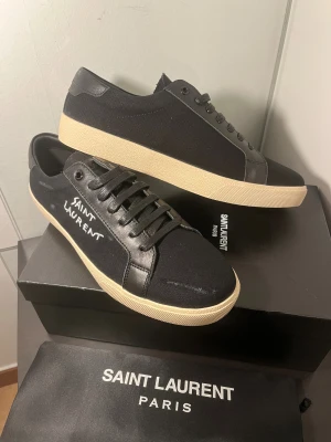 Saint Laurent skor  - Svarta sneakers från Saint Laurent med handskriven logga på sidan. Skorna har en rund tå, snörning och kontrasterande vit/beige platt sula. Materialet är en mix av canvas och skinn, vilket ger en stilren och modern vibe. Perfekta för dig som gillar exklusiva märken och minimalistisk design. Passar perfekt som julklapp eller liknande 