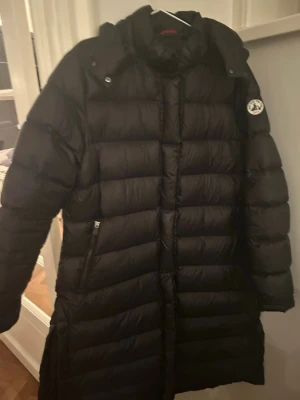 Svart lång dunjacka från JOTT - Säljer en svart lång dunjacka från JOTT med hög krage och huva. Jackan har quiltad design, dragkedjefickor och JOTT-logga på ärmen. Perfekt för kalla dagar och riktigt snygg till vintern.