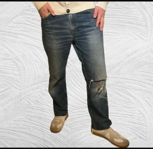  Archive Levi's 501 blå jeans med slitningar - Säljer ett par klassiska Levi's 521 jeans i blå tvätt med snygga slitningar och hål på ena knät. Jeansen har raka ben, fransade benslut och fem fickor. Perfekt för dig som gillar en avslappnad och vintage vibe. Byxorna är 34W 34L men är kapade till 95 cm. Hör av dig för minsta fundering!