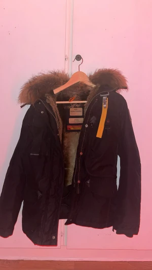 Svart Parajumpers jacka med äkta päls  - Svart parkas från Parajumpers i medium med fluffig brun päls runt huvan och insidan. Jackan har flera fickor, gul detaljrem och robust dragkedja. Perfekt för kalla dagar med sin varma och mjuka pälsfodring. Slim fit och snygg design.(OBS dragkedjan är sönder original pris 10.000)