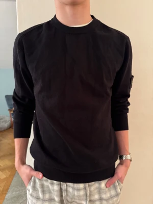 Svart Stone Island sweatshirt - Svart sweatshirt från Stone Island med klassisk rund hals och ribbade muddar. Tröjan har den ikoniska logotypen på vänster ärm och är tillverkad i mjuk bomull. Perfekt för dig som gillar stilrena och snygga streetwear-plagg.