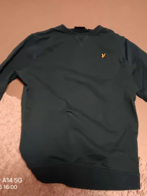 Mörkgrön sweatshirt från Lyle & Scott - Snygg mörkgrön sweatshirt från Lyle & Scott med klassisk gul logga broderad på bröstet. Tröjan har rund hals, långa ärmar och ribbade muddar vid ärmslut och nederkant. Perfekt för dig som gillar stilrena och enkla plagg.