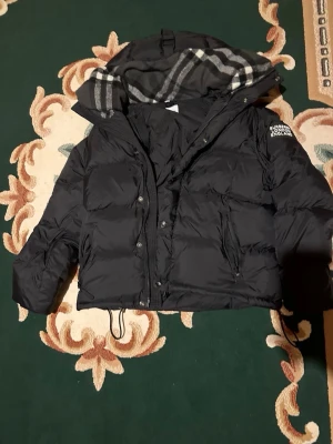 Svart Burberry pufferjacka med huva - Svart pufferjacka från Burberry med klassisk logga på ärmen och rutigt foder i huvan. Jackan har dragkedja, knappar och snörning nedtill. Flera fickor med dragkedja och en croppad passform som ger en cool vibe. Perfekt för kalla dagar.