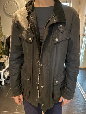 Svart Barbour waxjacka M - Barbour International Duke Wax Jacket är en klassiker för att hålla värmen på hojen, i stan eller på landet. Fyra utanpåliggande fickor. Duke är gjord i vaxad bomull som håller vätan ute. Quiltat foder. Patches på armbågarna gör den extra slitstark. Jackan är svart med svarta detaljer. Tvättas ej, rengörs med en våt trasa & vaxas om vid behov, lämna in din jacka hos Barbour-återförsäljare eller köp hem vax & gör det själv hemma Duke är en basvara från Barbour International,  nypris 4200:-