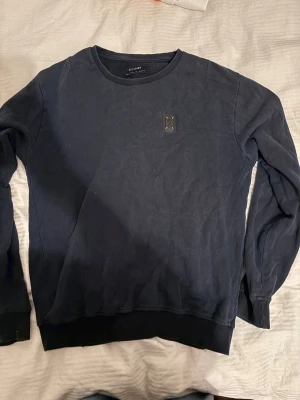 Mörkblå Vintage sweatshirt från Religion - Snygg mörkblå sweatshirt från Religion med rund hals och ribbade muddar vid ärmslut och nederkant. Tröjan har ett diskret patch på bröstet och sömdetaljer på ryggen. Perfekt för dig som gillar stilrena och enkla plagg. Vintage och svår å få tag på
