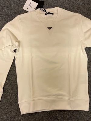 Vit sweatshirt från Prada - Snygg vit sweatshirt från Prada med klassisk rund hals och lång ärm. Tröjan har ett ikoniskt Prada-emblem i metall framtill vid halsen och ribbade muddar vid ärmslut och nederkant. Perfekt för dig som gillar stilrena designer och exklusiva märken.