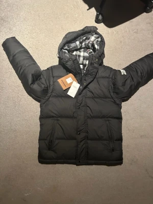 Svart pufferjacka från Burberry - Svart pufferjacka från Burberry med huva och rutig innerfoder i grått, vitt och rött. Jackan har dragkedja och knappar framtill, samt två sidofickor med dragkedja. Stor Burberry-logga på ärmen. Perfekt för kalla dagar och riktigt snygg stil.