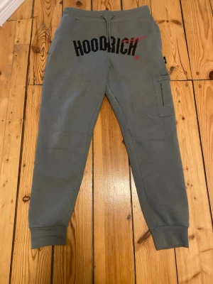 Grå Hoodrich Byxor - Hej, säljer ett par helt nya Hoodrich byxor då det var fel storlek. Byxorna är gråa med svart och röd logga framtill. Byxorna har snörning i midjan, ribbade muddar vid bensluten och en praktisk ficka på sidan. Perfekta för chill eller streetwear och tillverkade i mjukt bomullsmaterial.