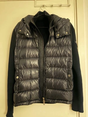 Svart pufferjacka från Moncler - Svart pufferjacka från Moncler med stickade ärmar och huva. Jackan har quiltad front i glansigt material, dragkedja och snygga detaljer med logotyp på ärmen. Perfekt för dig som vill ha en cool och varm jacka med streetkänsla.