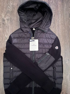 Svart pufferjacka från Moncler, strl L - Svart pufferjacka från Moncler med stickade ärmar och detaljer. Jackan har huva, dragkedja framtill och en ficka med dragkedja på ärmen. Kombinerar quiltad polyester med stickad material för en cool look. Moncler-logga på ärmen.