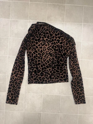 Leopardmönstrad oneshoulder topp Gina Tricot - Säljer en brun och svart mesh topp från Gina Tricot i storlek XS. Modellen är oneshoulder med lång ärm och har ett coolt leopardmönster över hela plagget. 