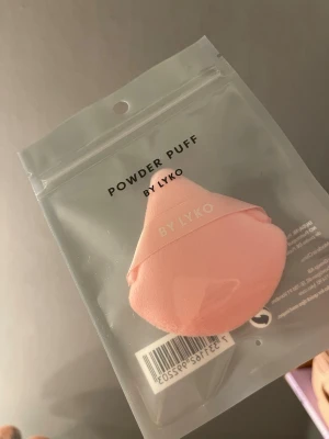 Powder Puff och Makeup Sponge från Lyko - Säljer ett set med en puder puff i ljusrosa och en makeupsvamp i lila från Lyko. Puffen har en mjuk yta och ett band för grepp, perfekt för att applicera puder. Svampen är droppformad och passar för foundation och concealer. Båda kommer i praktiska förpackningar. 