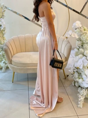 Champagnefärgad bal/bröllops klänning i satin - Säljer en elegant och figurnära långklänning i champagnefärgad satin. Klänningen är axelbandslös med en snygg slits på sidan som ger en extra touch. Materialet är glansigt och mjukt, perfekt för dig som vill sticka ut med en lyxig look. Den är figursydd hos en skräddare vid midjan och passar både XS och XXS. 