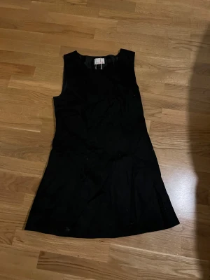 Svart ärmlös kortklänning Koola Anna - Svart ärmlös kortklänning från Koola Anna med diskret glittrig detalj i form av små svarta stenar som bildar loggan på nedre delen. Klänningen har en enkel, rak form och är tillverkad i ett mjukt syntetmaterial med slät yta. Perfekt för dig som gillar stilrena plagg med en twist.