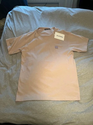 Beige t-shirt från Acne Studios - Snygg beige t-shirt från Acne Studios med klassisk rund hals och korta ärmar. På bröstet sitter Acne Studios ikoniska ansiktslogga i ljust lila. T-shirten är gjord i mjuk bomull och har en clean, minimalistisk vibe som funkar till allt.