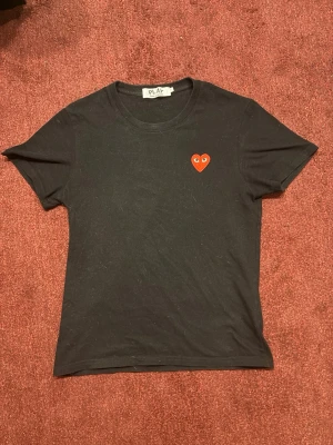 Svart t-shirt Comme des Garçons Play - Svart t-shirt från Comme des Garçons Play med klassisk röd hjärtlogga och knappt använd. Modellen är kortärmad och har en rund halsringning. Materialet är mjuk bomull som känns skön mot huden. Perfekt för dig som gillar stilrena och ikoniska plagg.