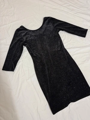 Glittrig svart klänning Sisters Point - Snygg svart kortklänning från Sisters Point med glittrigt tyg och rundad hals. Klänningen har trekvartsärm och en tight passform som framhäver figuren. Perfekt för dig som vill sticka ut med lite extra glitter. Materialet känns mjukt och stretchigt.