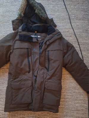 Brun parkajacka från Everest med fuskpäls - Brun parkajacka från Everest med huva och fuskpäls, flera praktiska fickor med dragkedja och stora framfickor. Jackan har lång ärm och är perfekt för kalla dagar. Materialet är polyester och syntet, och jackan har en klassisk parkamodell.