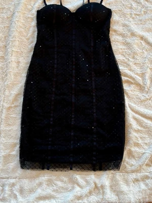 Svart glittrig kort klänning från NAKD - Supersnygg svart kort klänning från NAKD med glittriga paljetter och meshdetaljer. Klänningen har figurnära passform, kupor upptill och justerbara axelband. Perfekt för dig som vill sticka ut med lite extra glam. Materialet är polyester och mesh. Angivet storlek är 40. Men måtten överensstämmer med storlek 34, xs
