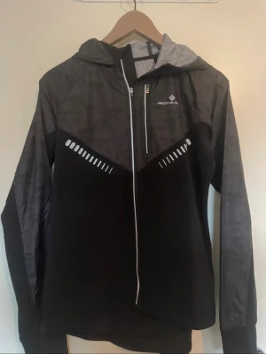 Svart träningsjacka  - En enkel träningsjacka från Ronhill i svart och grått med reflexdetaljer på bröstet och ryggen. Det mörkgråa materialet upptill är tunnare och det svarta nertill är lite tjockare. På sista bilden ser man även att det finns ett litet hål man kan sticka tummen i.  Kontakta privat vid intresse!💕