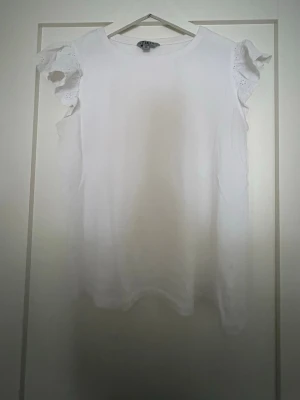 Vit blus med volangärmar från TIKI - Supersöt vit blus från TIKI med volangärmar i broderad spets. Blusen är ärmlös med rund halsringning och har en luftig passform. Perfekt till jeans eller kjol för en fräsch look.