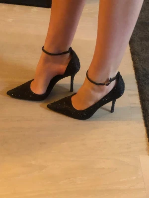 Svarta glittriga klackar  - Säljer ett par helt oanvända svarta klackar med glittriga detaljer och smal hög klack. Skorna har spetsig tå och en tunn rem runt ankeln med spänne. Bekväma att gå i! Nypris ca 500kr. 