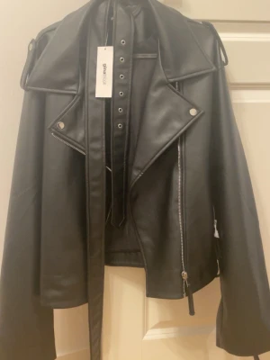 Stilig läderjacka -  OANVÄND! Svart skinnjacka (Läderimitation) Nytt skick! Svart, stilren jacka med bälte och dragkedja. Helt ny, aldrig använd – lapparna sitter kvar! Storlek L. Passa på!   