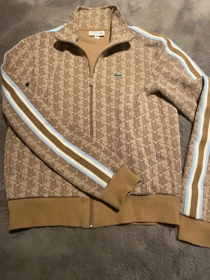 Beige mönstrad ziptröja Lacoste S - Säljer en beige ziptröja från Lacoste med brunt geometriskt mönster och blå/vita ränder längs ärmarna.  Tröjan är i mycket bra skick, hör av dig vid minsta fundering, priset är inte hugget i sten 🙌