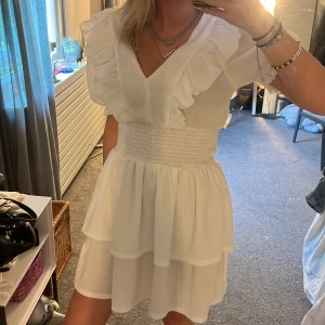 Vit volangklänning  - Supersöt vit kortklänning från Gina Tricot med v-ringning, volangdetaljer och puffärmar. Smockad midja och dubbla lager i kjolen ger en flowy känsla. Perfekt för sommarens alla tillfällen, riktigt fräsch och trendig look.