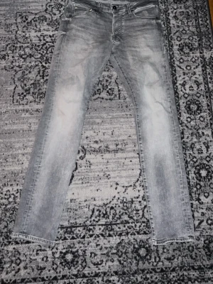 Grå jeans från Jack & Jones, 30/32 - Snygga grå jeans från Jack & Jones i storlek 30/32. Modellen har klassisk femficksdesign, raka ben och subtila slitningar för en cool look. Jeansen är tillverkade i bomullsmaterial och har en normal passform. Perfekt för dig som gillar stilrena och moderna jeans.