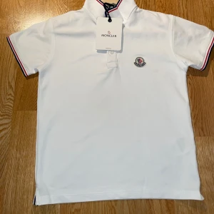 Vit monclear t-shirt  - Snygg vit pikétröja som är märket monclear  med klassisk krage och korta armar. Tröjan har rödblå ränder på krage och armslut samt Moncler-logga broderad på bröstet. Tillverkad i mjuk bomull som känns skön mot huden. Perfekt för dig som gillar stilrena märkesplagg.