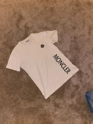 Vit Moncler t-shirt med logga - Säljer en clean vit t-shirt från Moncler i bomull, med stor svart Moncler-tryck på framsidan och en broderad logga på bröstet. Klassisk rund hals och korta ärmar, perfekt för dig som gillar stilrena designer och vill ha något som sticker ut.