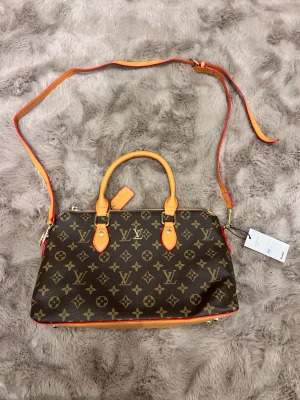 Louis Vuitton monogram axelväska - Säljer en snygg axelväska från Louis Vuitton med klassiskt monogrammönster i brunt och beige. Väskan har både handtag och justerbar axelrem i ljusbrunt skinn med röda detaljer. Guldiga metalldetaljer och dragkedja upptill. Perfekt storlek för dina essentials.
