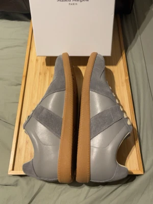 Maison Margiela grå sneakers - Maison Margiela sneakers i ljusgrå mocka och skinn med beige snörning och gummisula. Klassisk rund tå och platt sula, stilren design med kontrasterande material och subtil branding på plösen. Perfekt för dig som gillar minimalistisk och clean look.