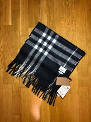 Svart rutig Burberry scarf i kashmir - Snygg svart halsduk från Burberry med klassiskt rutigt mönster i vitt och grått. Scarfen är tillverkad i 100% kashmir och har fransar längs nederkanten. Perfekt för dig som vill ha en lyxig och tidlös look med premiumkänsla. Använd en gång och säljer då färgen inte var min stil.